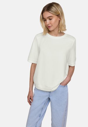 Frau mit schulterlangem blondem Haar, die ein lockeres weißes kurzärmeliges Oberteil und hellblaue Jeans trägt und mit einer Hand in der Tasche steht.