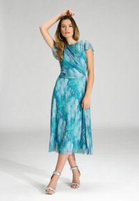 Niebieska i zielona sukienka w abstrakcyjny wzór z krótkimi bufkami, dopasowaną górą i zwiewną spódnicą, w połączeniu z srebrnymi sandałami na obcasie.