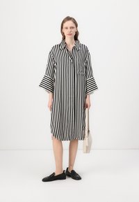 Mekko, jossa on mustavalkoiset raidat, kaulus, lyhyet hihat, sivutasku ja pyöristetty helma, yhdistettynä mustiin tasapohjaisiin kenkiin ja vaaleaan laukkuun.