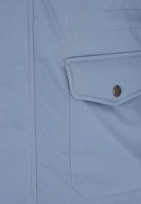 Giacca di tessuto azzurro chiaro con tasca sul petto con patta e chiusura a bottone, e cuciture visibili lungo le cuciture. Texture liscia.