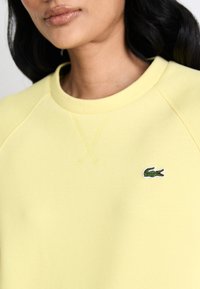 Sweatshirt amarelo de algodão com decote redondo, apresentando um pequeno logótipo de crocodilo bordado em verde no lado esquerdo do peito. Tecido texturizado.