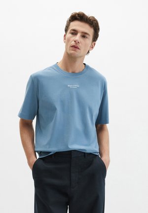 Jonge man met krullend haar, gekleed in een blauw Marc O’Polo T-shirt en een zwarte broek, met zijn handen in zijn zakken, kijkt iets opzij.
