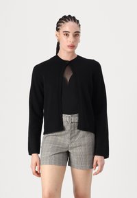 Μαύρο καρντιγκάν με στρογγυλή λαιμόκοψη, ανοιχτό μπροστά και μακριά μανίκια, συνδυασμένο με καρό σορτς που διαθέτει ζώνη στη μέση.