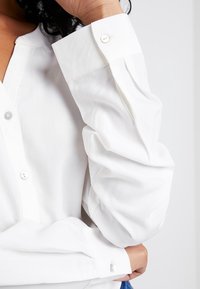 Chemise blanche en tissu léger, dotée d'un col boutonné, de manches longues bouffantes et de petits boutons lisses aux poignets.