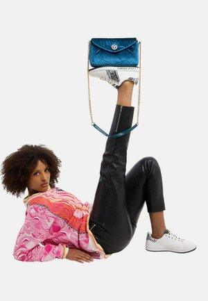 Frau liegt auf dem Rücken, trägt schwarze Lederhose und pinken Pullover, hält eine blaue Handtasche balancierend auf einem senkrecht erhobenen, mit Sneakers bekleideten Fuß.