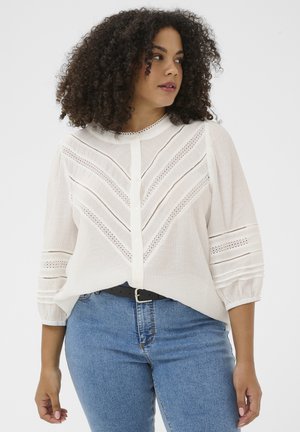 Femme en blouse blanche boutonnée avec des détails en dentelle et des manches trois-quarts, associée à un jean bleu et une ceinture noire, regardant vers la droite.