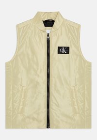 Gai dzeltens bezpaliku veste ar augstu apkakli, priekša ar rāvējslēdzēju, melno rāvējslēdzēju un logotipa plāksteri kreisajā krūtī. Gluda, spīdīga tekstūra.