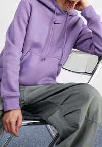 Lila hoodie med en känguruficka och svart Nike-logotyp. Matchad med gröna cargobyxor. Modell sitter på en kromstol.