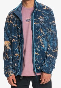 Chaqueta de forro polar con un patrón jaspeado en azul y naranja, con bolsillos con cremallera y un cuello alto. Combinada con una camiseta rosa claro debajo.