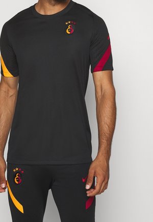 Uomo che indossa un abbigliamento sportivo nero con accenti rossi e gialli e il logo del club di calcio Galatasaray sul petto e sui pantaloni.