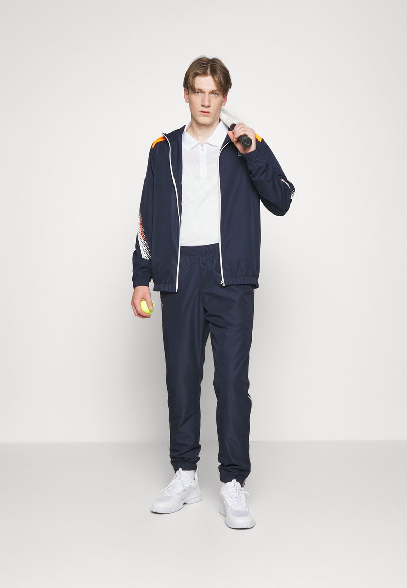 Survêtement bleu marine avec des accents orange, veste zippée, pantalon à taille élastique et baskets blanches. Tenant une raquette de tennis et une balle.