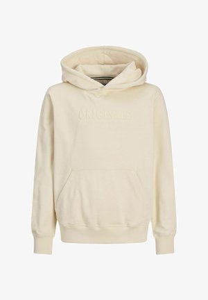 Osoba ubrana w kremową bluzę z kapturem i kieszenią z przodu, jasnoniebieskie szerokie dżinsy i białe trampki, stojąca na tle jednolitej białej ściany.