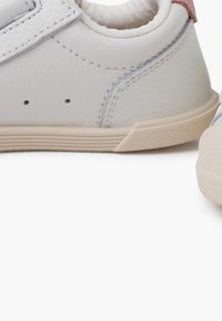 Sneakers sintetici bianchi con una finitura testurizzata, cuciture chiare e suole in gomma beige. Presentano un piccolo accento rosa sul tallone.