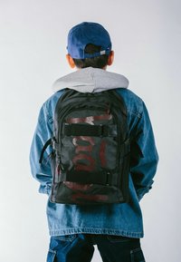 Zaino nero con dettagli in rete rossa e fibbie, caratterizzato da un design strutturato. Indossato da una persona con una giacca di jeans e un felpa grigia.