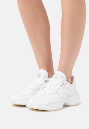 Ben iført hvide Adidas-sneakers med snørebånd, stående på en hvid overflade.