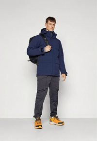 Marineblå isolert jakke med hette, kombinert med mørkegrå cargobukser og oransje tursko, med en svart ryggsekk.