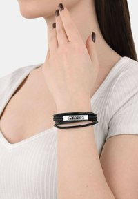 Braccialetto in pelle nera con un design intrecciato e una placca in metallo color argento incisa con "SCORPIONS." Indossato su un polso.