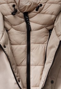 Veste matelassée beige avec fermeture éclair noire et boutons à cordon sur la capuche, montrant des coutures détaillées et la texture du tissu.