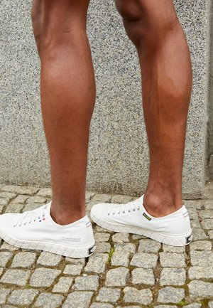 Dos piernas con zapatillas de lona blancas de corte bajo, de pie sobre un pavimento de piedra frente a una pared de piedra gris.