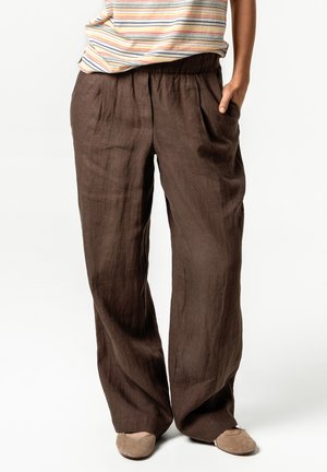 Brown loose linen pants with elastic waist worn with beige flats and a striped tucked-in shirt, hand in pocket.