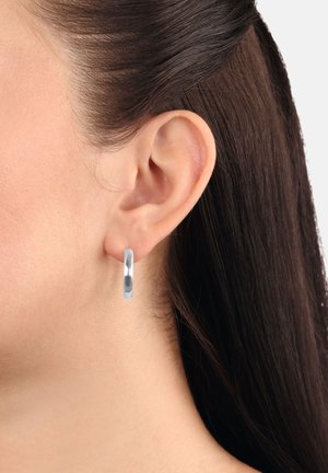 Orecchio destro di una donna con capelli scuri dietro, che indossa un orecchino a cerchio in argento lucido su sfondo bianco.