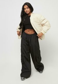 Kvinne med langt mørkt hår, iført en beige bomberjakke, svart kort topp, vide svarte cargo-bukser og svart-hvite sneakers.