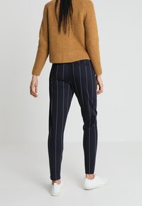 Navy trousers with white pinstripes, tapered design, and pockets; paired with a textured mustard sweater and white sneakers.