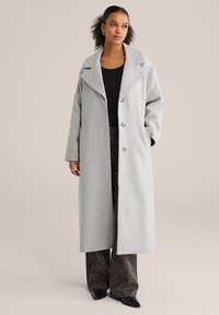 Palton din lână gri deschis cu rever larg, nasturi cu două rânduri și buzunare laterale. Asortat cu pantaloni întunecați cu model și pantofi negri.