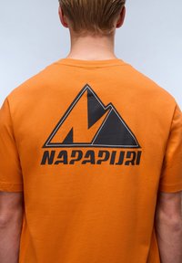 Orange bomulds t-shirt med et sort bjergdesign og "NAPAPIJRI" tekst på ryggen. Korte ærmer og klassisk rund halsudskæring.