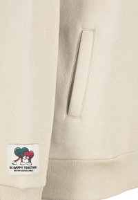 Beige sweatstof met een zijzak, met een patch van cartoonhartjes en de tekst "WEES GELUKKIG SAMEN MET POSITIEVE VIBES."