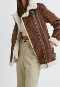 Chaqueta de ante sintético marrón con forro de borreguito blanco, cierre con cremallera en la parte delantera y detalles en cuero marrón. Presenta bolsillos laterales y una textura suave.