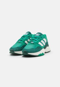 Un paio di sneakers Adidas verdi con strisce bianche, suole intermedie bianche e suole in gomma verde scuro, angolate leggermente a destra su uno sfondo bianco.