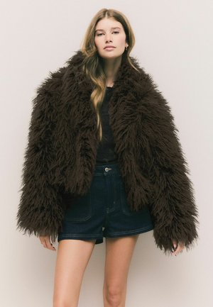 Brun faux-fur jakke med en fluffy tekstur, oversized form og brede ærmer, parret med mørke denim shorts og en tætsiddende top.