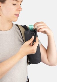 Schwarze Stofftasche für eine Wasserflasche mit Kordelzugverschluss, befestigt an einem hellen Schultergurt. Eine Flasche mit grünem Verschluss wird gerade hineinsteckt.