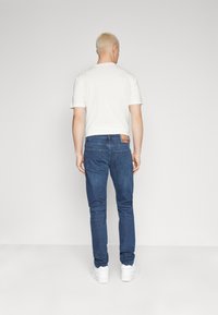 Jean de mezclilla azul oscuro con un corte ajustado, que presenta una etiqueta de parche de cuero en la parte trasera y sutiles detalles de costura, combinado con una camiseta blanca lisa.