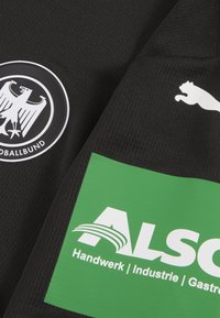 Must kandetussärk, millel on ümmargune must-valge Saksamaa jalgpalliliidu logo ja roheline ristkülikukujuline sponsorilogo valge tekstiga.