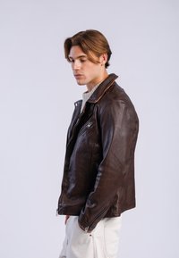 Veste en cuir marron de moto avec col montant, poches zippées et détails de couture discrets, portée sur un pull en tricot gris clair.