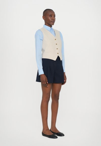 Beige liivi nappien kanssa, vaaleansininen nappipaita, tummansiniset laskostetut hameet ja mustat ballerinat, joita kantaa seisova henkilö.