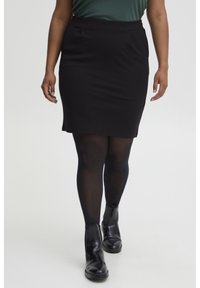 Zwarte aansluitende kokerrok met twee vo pockets, een gladde textuur en een lengte tot halverwege de dij. Gemaakt met zwarte panty’s en enkellaarsjes.