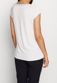 Weißes T-Shirt mit kurzen Ärmeln, Rundhalsausschnitt und glatter Textur, kombiniert mit schwarzen Hosen mit seitlichen Streifen. Keine sichtbaren Muster oder Akzente.