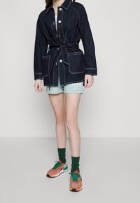 Denim jacket with a belted waist, contrasting white stitching, large front pockets, and a collared neck. Paired with light green shorts and colorful sneakers.