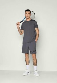Hall mõtlemisvõime juhtiv hall sporditootega T-särk ja lühikesed püksid, millel on suur must "BORG" logo. Mudel hoiab tennisereketit, kandes valgeid jalatseid ja sokke.