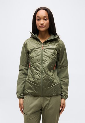 Žena stojící čelem vpřed, oblečená v olivově zelené prošívané bundě se zipem a odpovídajících kalhotách na jednobarevném bílém pozadí.