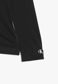 Svart långärmad skjorta med en jämn yta, med en vit logotyp "cK" på ärmsluten och en detaljerad slits på sidan.