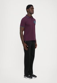 Νέος άνδρας στέκεται πλάγια φορώντας ένα μωβ πόλο με μπλε λεπτομέρειες, μαύρο παντελόνι και sneakers μαύρου και άσπρου χρώματος σε λευκό φόντο.