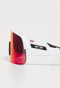 Gafas de sol deportivas blancas con grandes lentes rojos de degradado, acentos negros texturizados en las patillas y un logo Prizm en la lente y en el lateral.