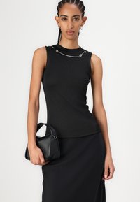 Zwarte geribde tanktop met zilveren kettingdetails op de schouders, gecombineerd met een zwarte handtas. De stof is rekbaar en aansluitend.