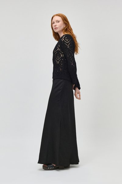 Femme aux longs cheveux roux portant un pull noir à maille ajourée, une longue jupe noire et des sandales compensées noires, debout devant un fond clair.
