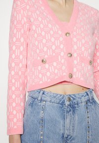 Różowy, krótki kardigan z teksturowanym wzorem i dekoltem w serek, ozdobiony metalowymi guzikami i przednimi kieszeniami, zestawiony z jasnoniebieskimi dżinsami.