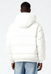 Giacca imbottita bianca con cappuccio, caratterizzata da un design trapuntato, polsini a coste e una vestibilità morbida. Indossata sopra jeans azzurri.